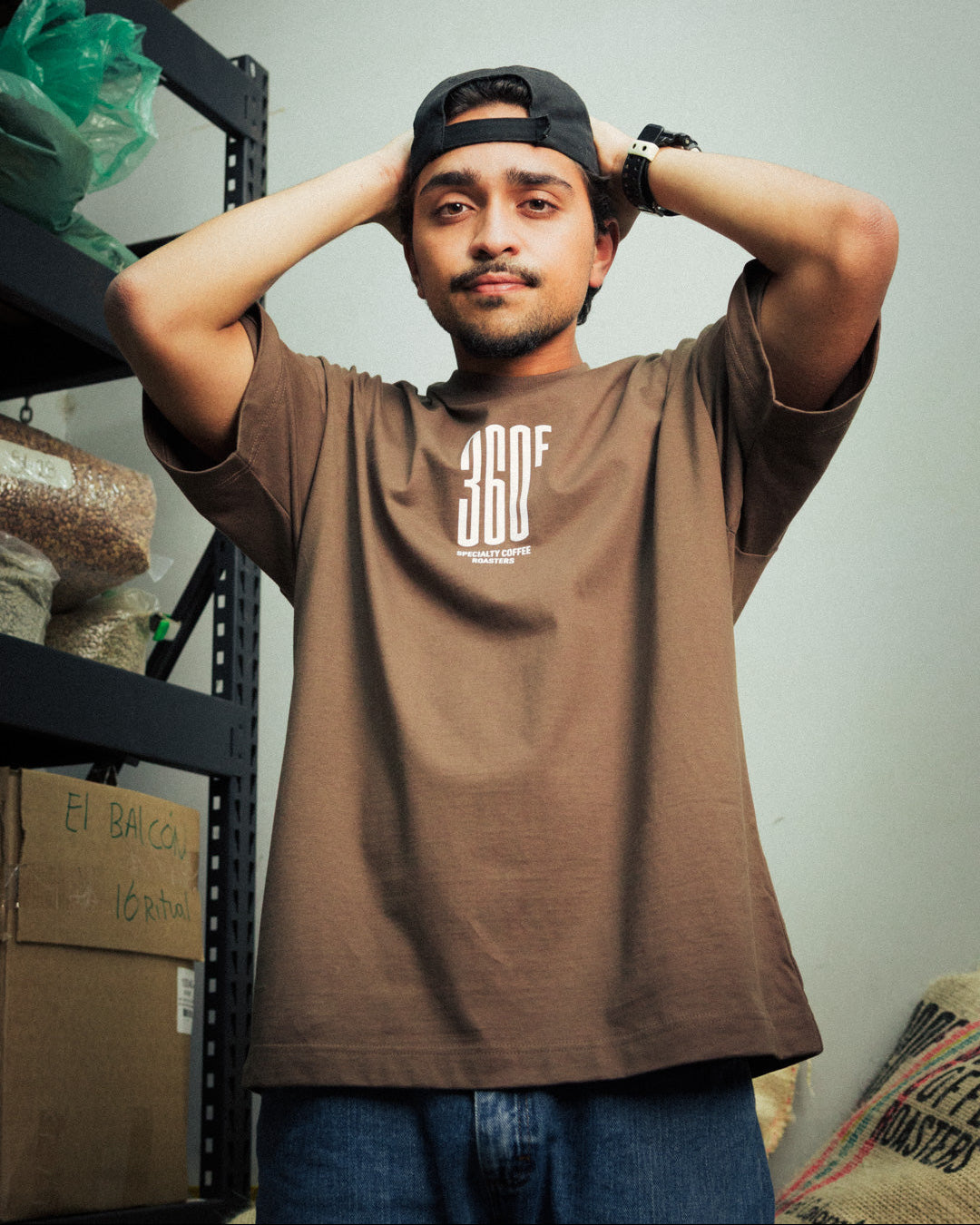 Person wearing a brown t-shirt with a logo, standing in a room with shelves and boxes.
