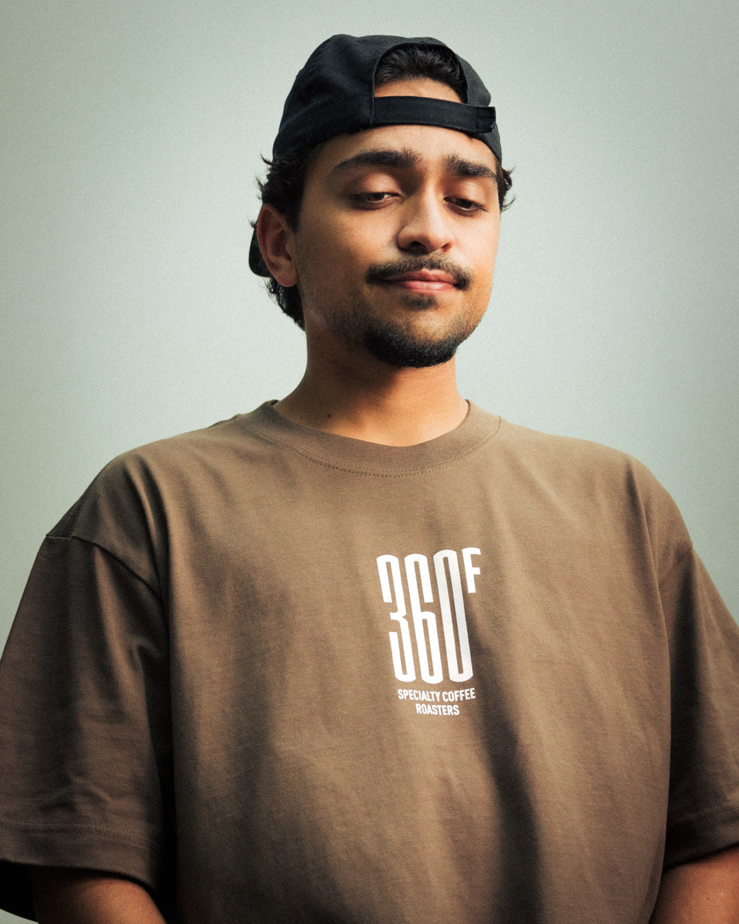 Man wearing a brown t-shirt with '360F' logo and a black cap on a light gray background