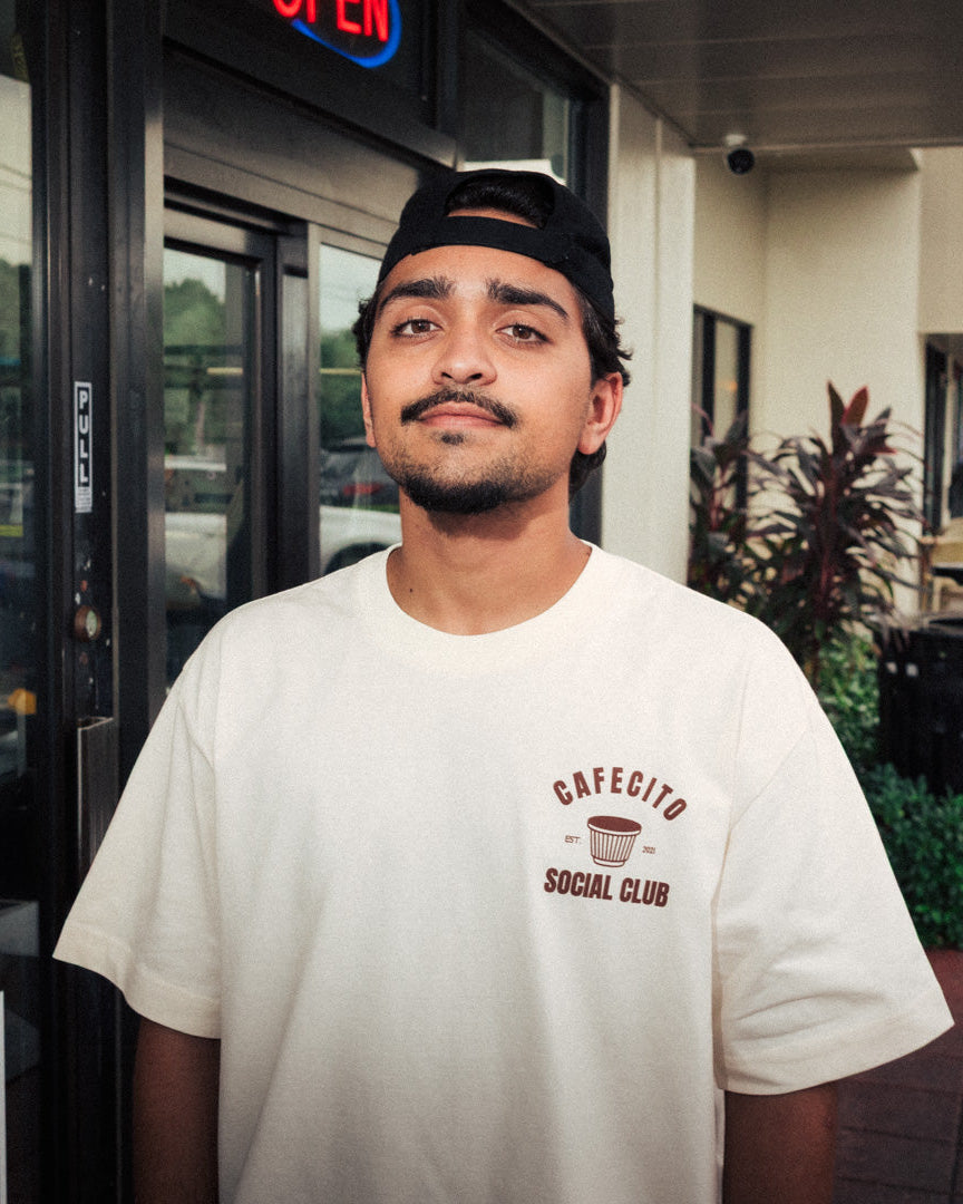 Man wearing a white t-shirt with 'Cafecito Social Club' logo standing outside a building.