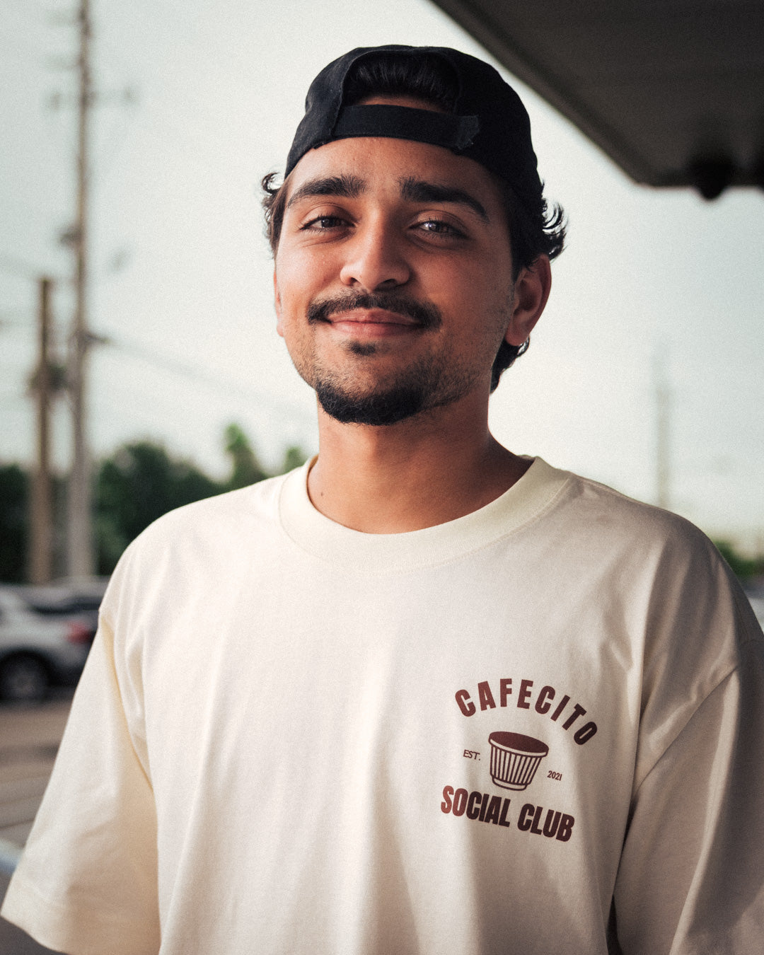 Man wearing a white t-shirt with 'Cafecito Social Club' logo, standing outdoors.